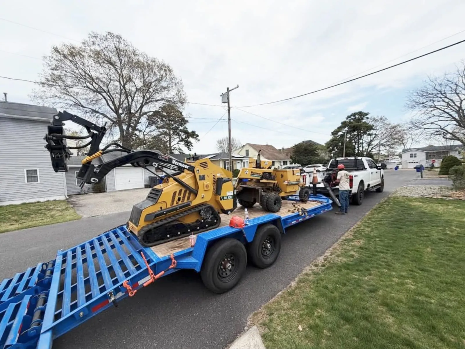 NJ Roots crew truck towing a trailer loaded with a mini skid-steer and stump grinder through a Jersey Shore neighborhood