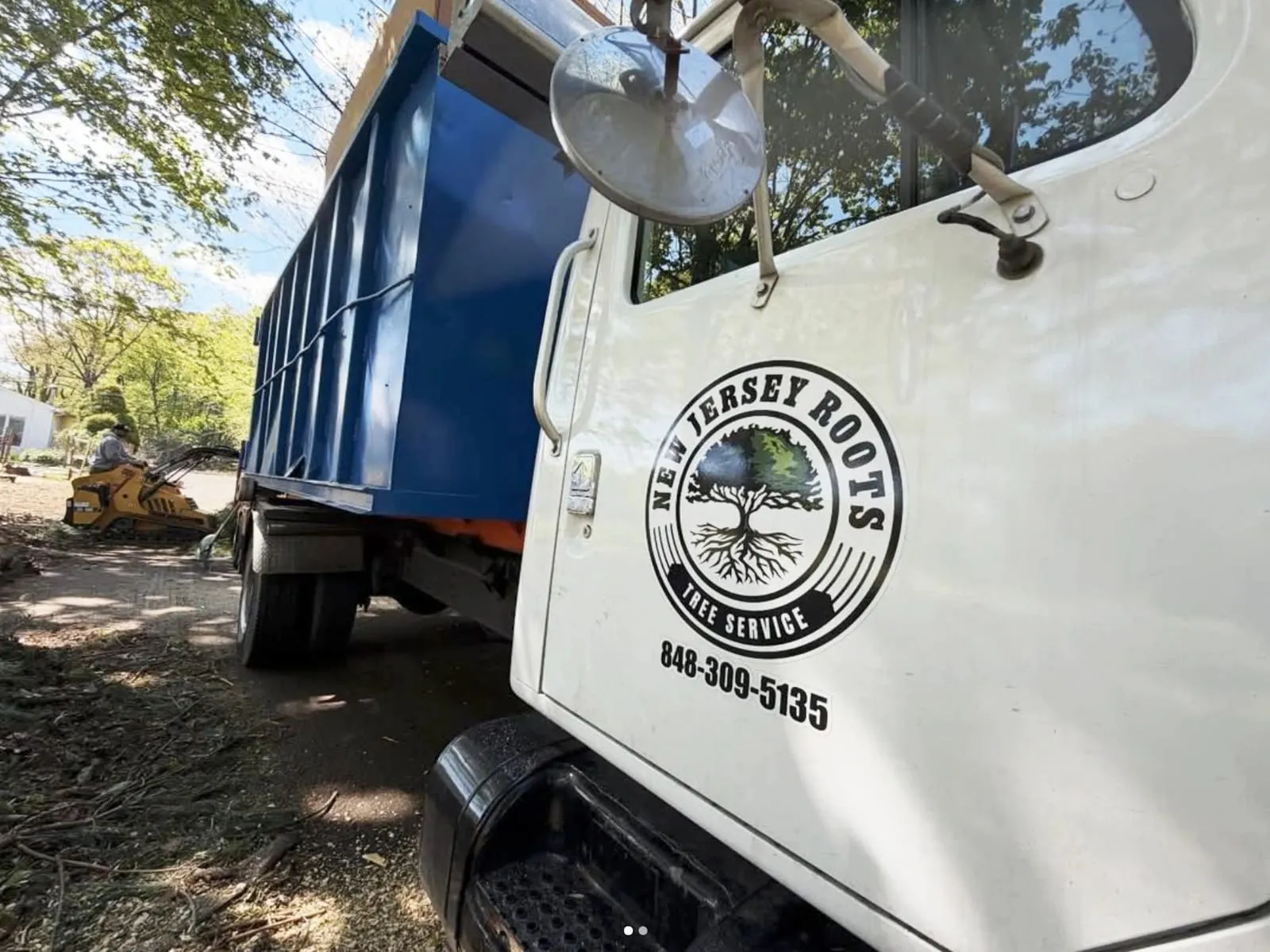 NJ Roots chip truck with the New Jersey Roots Tree Service badge and phone number on the door, with a loader working behind it on a job site